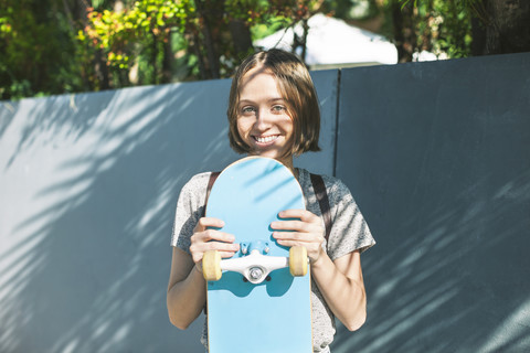 Porträt einer lächelnden jungen Skateboarderin, die ihr Skateboard hält, lizenzfreies Stockfoto