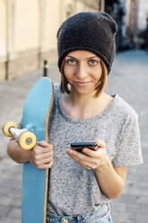 Porträt einer lächelnden jungen Skateboarderin mit Smartphone und Skateboard - EBSF000290