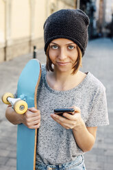 Portrait of smiling young female skate boarder holding smartphone and skateboard - EBSF000290