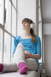 Porträt einer jungen Frau, die auf dem Fensterbrett in einem Büro sitzt und mit Kopfhörern Musik hört - RBF001690