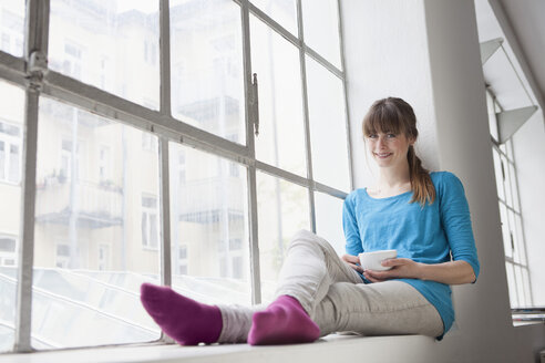 Portrait of smiling young woman with cup of coffee sitting on window sill in an office - RBF001691