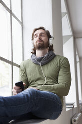 Porträt eines jungen Mannes, der auf der Fensterbank in einem Büro sitzt und mit Kopfhörern Musik hört - RBF001693