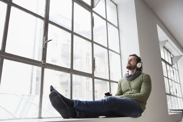 Portrait of young man sitting on window sill in an office listening music with headphones - RBF001694