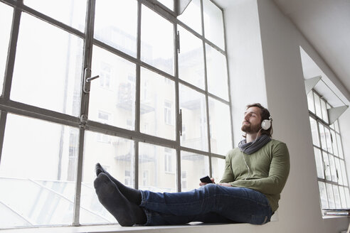 Porträt eines jungen Mannes, der auf der Fensterbank in einem Büro sitzt und mit Kopfhörern Musik hört - RBF001694