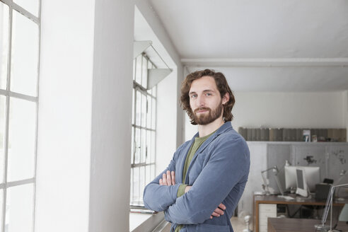 Porträt eines jungen Mannes mit verschränkten Armen, der in einem Büro steht - RBF001699