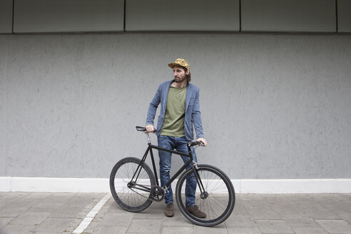 Deutschland, Bayern, München, junger Mann mit Basecap steht mit seinem Rennrad vor einer Mauer - RBF001705