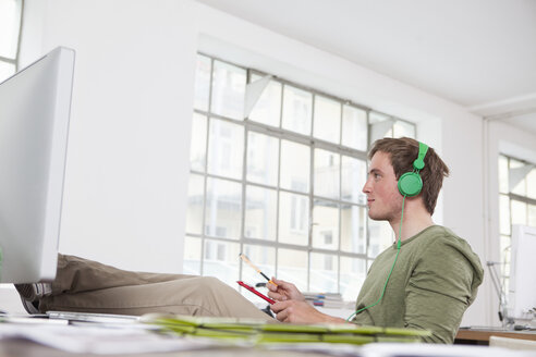 Young man relaxing with feet on his desk and hearing music with headphones in the office - RBF001713