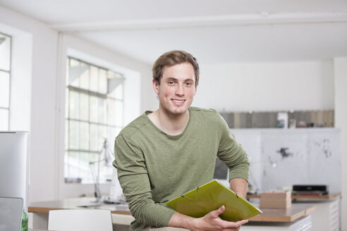Portrait of smiling young man sitting on his desk in the office - RBF001787