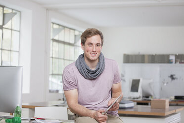 Porträt eines lächelnden jungen Mannes, der an seinem Schreibtisch im Büro sitzt und ein digitales Tablet hält - RBF001788