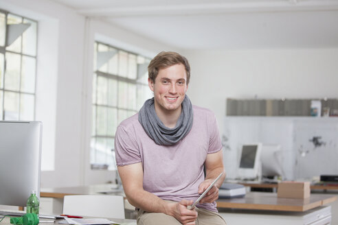 Portrait of smiling young man sitting on his desk in the office holding digital tablet - RBF001788