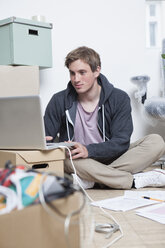 Porträt eines jungen Mannes, der in einem Büro zwischen Kartons auf dem Boden sitzt und sein Notebook benutzt - RBF001714
