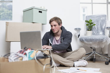 Porträt eines jungen Mannes, der in einem Büro zwischen Kartons auf dem Boden sitzt und sein Notebook benutzt - RBF001715