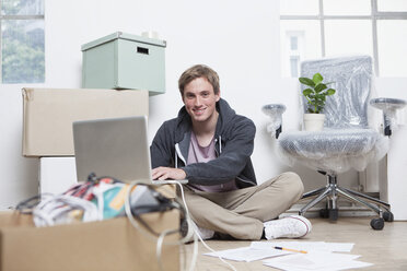 Porträt eines jungen Mannes, der in einem Büro zwischen Kartons auf dem Boden sitzt und sein Notebook benutzt - RBF001716
