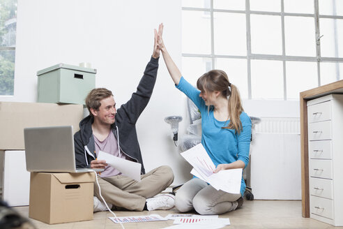 Two colleagues sitting on ground between cardboard boxes in an office giving high five - RBF001718