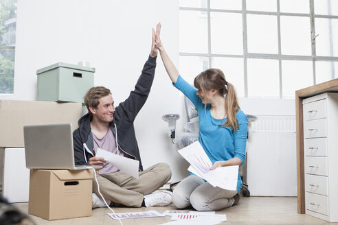 Zwei Kollegen sitzen auf dem Boden zwischen Kartons in einem Büro und geben sich ein High Five - RBF001718