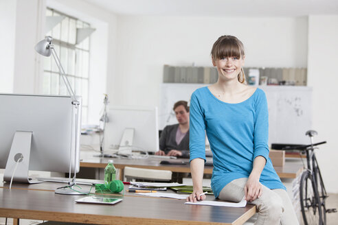 Portrait of smiling young woman sitting on her desk in an office - RBF001725