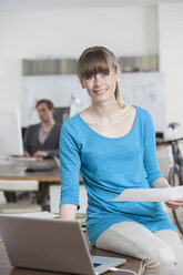 Porträt einer lächelnden jungen Frau, die an ihrem Schreibtisch in einem Büro sitzt - RBF001727