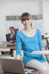 Portrait of smiling young woman sitting on her desk in an office - RBF001727