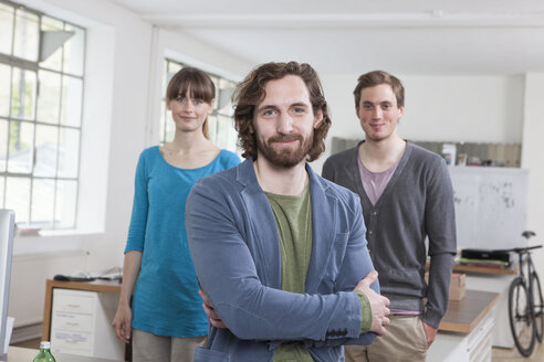 Portrait of young man with his two colleagues in the background standing in a creative office - RBF001741