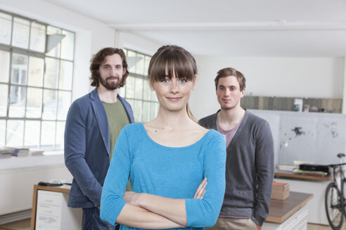 Portrait of young woman with her two colleagues in the background standing in a creative office - RBF001742