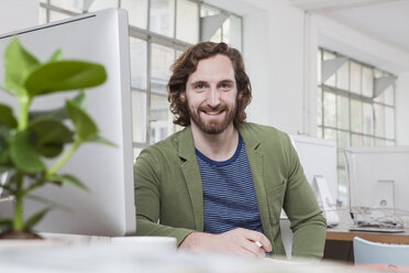 Portrait of young man sitting at his desk in a creative office - RBF001760