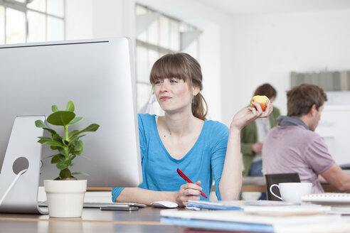 Portrait of young woman eating an apple at her desk in a creative office - RBF001795