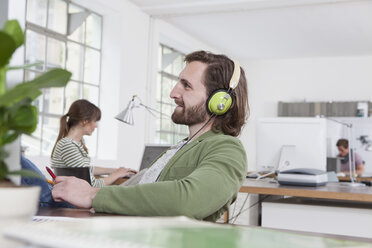 Junger Mann sitzt an seinem Schreibtisch in einem kreativen Büro und hört Musik mit Kopfhörern - RBF001784