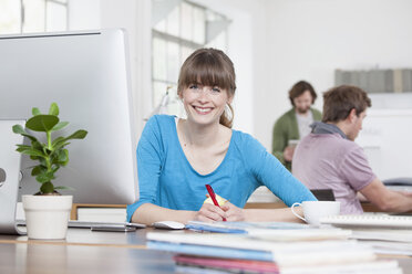 Porträt einer lächelnden jungen Frau, die an ihrem Schreibtisch in einem kreativen Büro sitzt - RBF001785