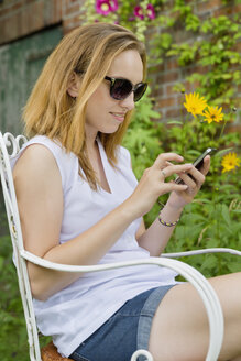 Junge Frau mit Sonnenbrille sitzt in einem Garten und benutzt ihr Smartphone - SEF000805