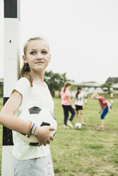 Porträt eines lächelnden Mädchens mit Fußball, das am Torpfosten lehnt - UUF001553