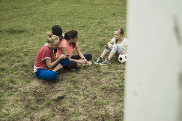 Four female teenage friends sitting on soccer field using their smartphones - UUF001580
