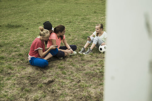 Vier Freundinnen im Teenageralter sitzen auf einem Fußballfeld und benutzen ihre Smartphones - UUF001580