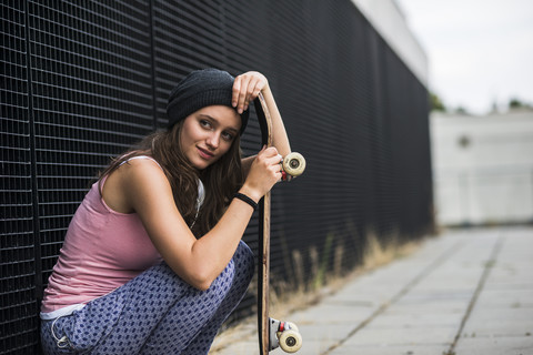 Smiling teenage girl with skateboard crouching in front of a black facade stock photo