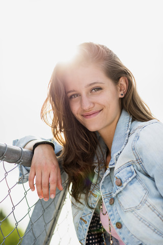 Porträt eines lächelnden Teenagers mit Jeansjacke, der sich an einen Zaun lehnt, lizenzfreies Stockfoto