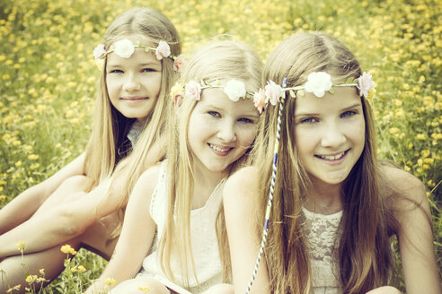 Portrait of three girls wearing floral wreaths sitting on a flower meadow - GDF000391