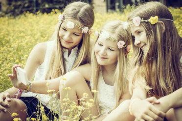 Portrait of three girls wearing floral wreaths taking a selfie on a flower meadow - GDF000390