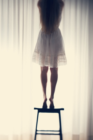 Junge Frau steht auf einem Hocker vor einem weißen Vorhang, Rückansicht, lizenzfreies Stockfoto