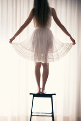 Junge Frau steht auf einem Hocker und hält den Saum ihres Rockes vor einem weißen Vorhang, Rückansicht - BRF000580