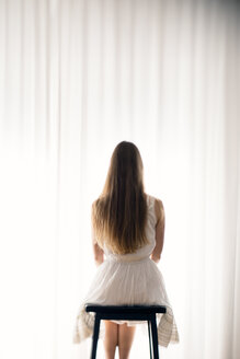 Junge Frau sitzt auf einem Hocker vor einem weißen Vorhang, Rückansicht - BRF000582