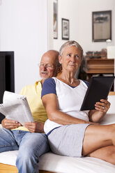 Senior couple sitting on couch reading newspaper using digital tablet - JUNF000014