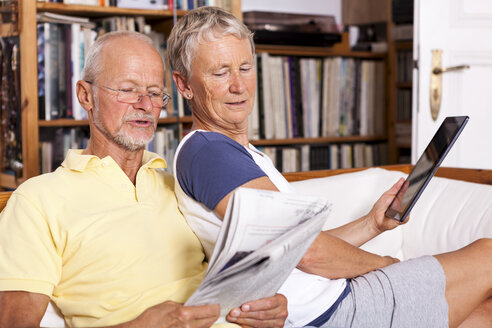 Senior couple sitting on couch reading newspaper using digital tablet - JUNF000015