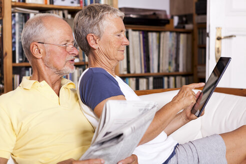 Älteres Paar sitzt mit Zeitung auf der Couch und benutzt ein digitales Tablet - JUNF000009