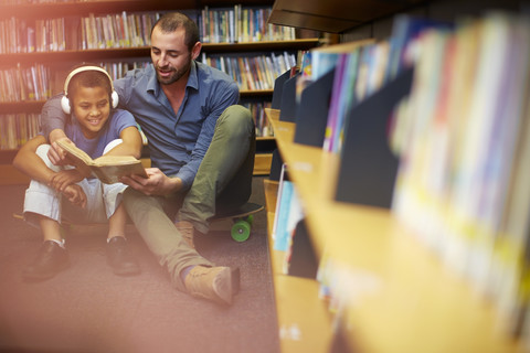 Mann liest einem Jungen mit Kopfhörern in der Bibliothek ein Buch vor, lizenzfreies Stockfoto