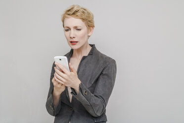 Porträt einer verzweifelten blonden Frau, die auf ihr Smartphone schaut, vor einem grauen Hintergrund - TCF004235