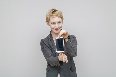 Porträt einer lächelnden blonden Frau, die ihr Smartphone vor einem grauen Hintergrund zeigt - TCF004242