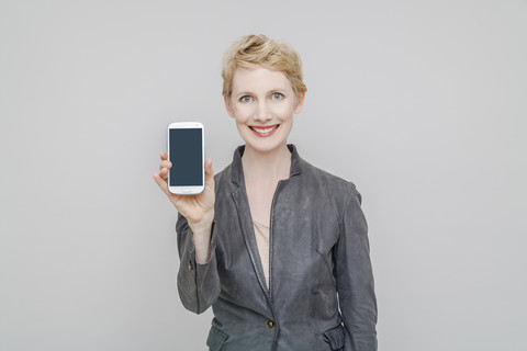 Porträt einer lächelnden blonden Frau, die ihr Smartphone vor einem grauen Hintergrund zeigt, lizenzfreies Stockfoto