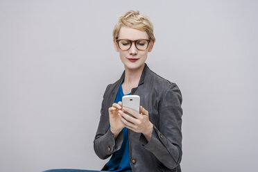 Porträt einer lächelnden blonden Frau, die ein Smartphone vor einem grauen Hintergrund benutzt - TCF004271