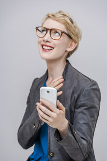 Porträt einer lächelnden blonden Frau mit ihrem Smartphone vor einem grauen Hintergrund - TCF004273
