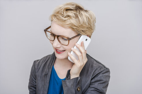 Porträt einer lächelnden blonden Frau, die mit einem Smartphone telefoniert, vor einem grauen Hintergrund - TCF004280