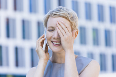 Lächelnde blonde Frau, die mit einem Smartphone telefoniert und ein Auge mit der Hand bedeckt - TCF004296